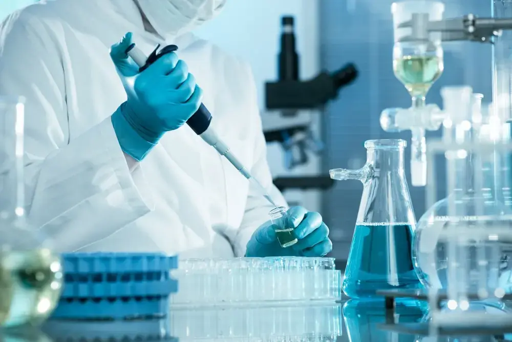Crime lab scientist in protective gear pipetting an evidence sample beside a microscope and laboratory flasks